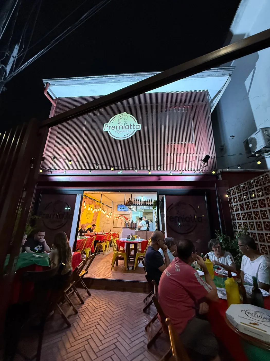 Fachada da Premiatta Pizzaria à noite, com clientes nas mesas da varanda